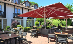 Außenbereich eines Restaurants mit roten Sonnenschirmen, Tischen und Stühlen vor einem Gebäude mit Blumen und gepflegter Bepflanzung.