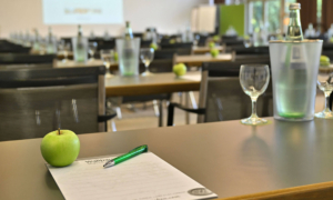 Im Vordergrund liegt auf einem Konferenztisch ein grüner Apfel neben einem Block und einem grünen Kugelschreiber. Dahinter stehen mehrere Tische mit Gläsern und Wasserflaschen. Im Hintergrund sind große Fenster mit Blick ins Grüne und eine Leinwand zu sehen. Die Szene wirkt wie ein Tagungsraum, der für eine Veranstaltung vorbereitet ist.