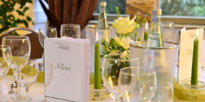 Landhotel-Schnuck-Bankett-0318-038_b5aebfcf51 Festlich gedeckter Tisch mit Menükarte, Kerzen, Gläsern, Mineralwasser und Blumendekoration.