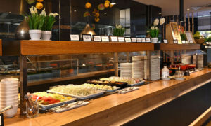 Buffet-Theke in einem Restaurant mit Holzoberfläche, gefüllt mit frischem Obst, Salaten und kleinen Gläsern; darüber Schilder mit Speisenamen und dekorative Pflanzen.