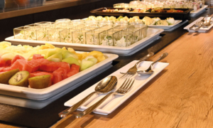 Nahaufnahme eines Buffets mit frischem Obst wie Kiwi, Melone und Wassermelone sowie kleinen Gläsern mit Kräutercreme; daneben liegen sauber arrangierte Bestecke auf weißen Tellern.