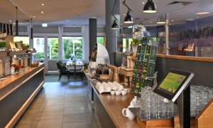 Moderner Frühstücksbereich mit Holztheke, weißen Tassen, Gläsern und Teeboxen; im Hintergrund Tische und Stühle vor großen Fenstern mit Blick ins Grüne.