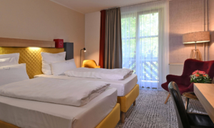 Ein helles Hotelzimmer mit zwei Einzelbetten, einem gelben Kopfteil, einer Sitzecke am Fenster und warmer Beleuchtung.