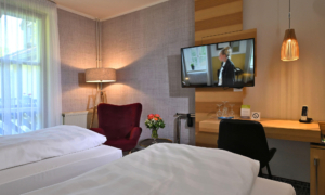 Modernes Hotelzimmer mit zwei Einzelbetten, einem Schreibtisch, einem roten Sessel und einem großen Fenster mit warmem Licht.