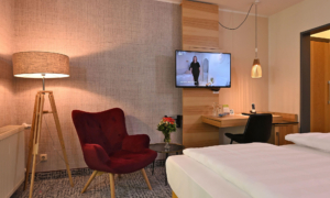 Modernes Hotelzimmer mit Bett im Vordergrund, rotem Sessel, Schreibtisch und warmem Licht.