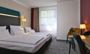 Bright hotel room with two single beds, colorful accents, a desk, and a seating area by the window.