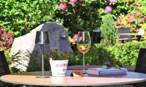 Holz­tisch im Garten mit kleiner Lampe, Blumentopf, Weinglas, Buch und Sonnenbrille vor grüner Bepflanzung.