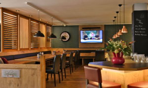 A warm bar scene featuring wooden furniture, dark leather armchairs, a TV on the wall, and a chalkboard beer menu.
