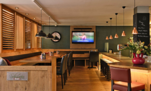Warme Bar­szene mit Holzmöbeln, dunklen Ledersesseln, TV an der Wand und beschrifteter Bier­tafel.