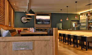 A warm bar scene featuring wooden furniture, dark leather armchairs, a TV on the wall, and a chalkboard beer menu.