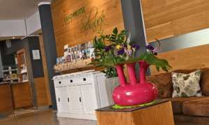 Helle Hotellobby mit Holzrückwand und grünem Schriftzug Landhotel Schnuck, davor ein weißes Prospektregal; rechts ein braunes Sofa mit Zierkissen und ein Holztisch mit pinker Vase und Blumen.