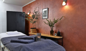 Ruhiger Behandlungsraum mit Massageliege, Kerze, warmem Licht und dekorierten Regalen an der Rückwand.