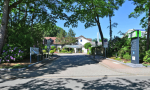 Außenansicht eines hellen Gebäudekomplexes mit rotem Dach, Glas­eingang und gepflegtem Gartenbereich.