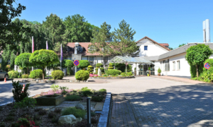 Außenansicht eines hellen Gebäudekomplexes mit rotem Dach, Glas­eingang und gepflegtem Gartenbereich.