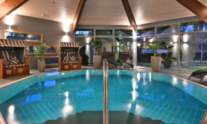 A modern indoor pool with clear blue water, sun loungers, wooden cabanas, and large plants in a bright setting.