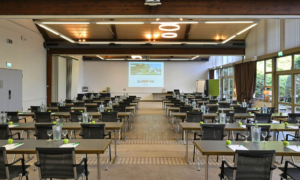 Großer heller Tagungsraum mit Reihen von Tischen und Stühlen, auf den Tischen stehen Wasserflaschen, Gläser und grüne Äpfel. Im Hintergrund an der weißen Wand befindet sich eine Beamerprojektion.