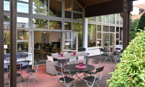 Überdachte Terrasse mit mehreren runden und rechteckigen Tischen, die mit Stühlen und Sitzgruppen aus Metall und Polyrattan ausgestattet sind. Auf den Tischen stehen dekorative Vasen mit rosa Blumen. Die Terrasse grenzt an ein Gebäude mit großen Fensterfronten, durch die ein heller Tagungsraum zu sehen ist.