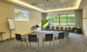 Heller Konferenzraum mit grauem Teppichboden und großen Fenstern, die Tageslicht hereinlassen. In der Raummitte steht ein Stuhlkreis aus schwarzen Kunststoffstühlen. An der linken Wand befindet sich eine eine projizierte Präsentation, daneben ein Flipchart mit handgeschriebenem Text. Im Hintergrund stehen ein Tisch mit technischem Equipment und eine grüne Trennwand. In der rechten Ecke sind zwei Sessel und ein kleiner Tisch zu sehen.