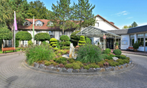 Außenansicht des Landhotel Schnuck mit weißem Fachwerkstil, roten Ziegeldächern und überdachtem Eingangsbereich. Ein gepflegter Vorgarten mit Ziergräsern, Formschnittgehölzen und einem dekorativen Steingarten begrüßt die Gäste.
